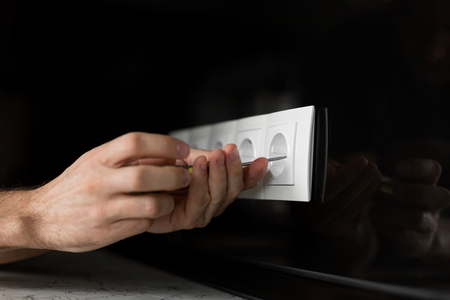 Close-up of an electrician's hand with a screwdriver disassembling a white electrical outlet on a black glass wall.の写真素材
