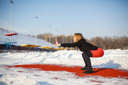 Young caucasian female blonde in red leggings stretching exercise on a red running track in a snowy stadium. fit and sports lifestyle.の写真素材