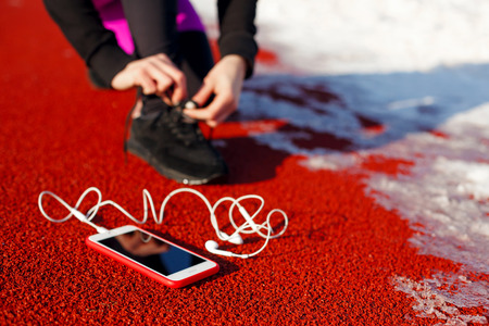 girl athlete in black sneakers, crouching on the red track for running. near is a phone with wired...