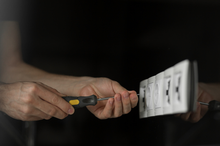 Close-up of an electrician's hand with a screwdriver disassembling a white electrical outlet on a black glass wall.の写真素材