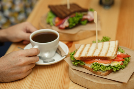 man takes a cup of coffee from a wooden table, on which lies a sandwich.の写真素材