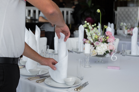 The waiter in the restaurant set up a table for guests. Waiter's hands are a white tablecloth, napkins, white dishes and a bouquet of flowers on the table in the frame.の写真素材