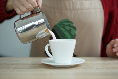 Barista pours milke from steel pot to white cup with coffee.