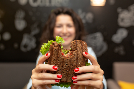 Closeup funny blurred protrait of young woman hold bitten sandwich by her two hands. Sandwich in focus. dark background.の写真素材