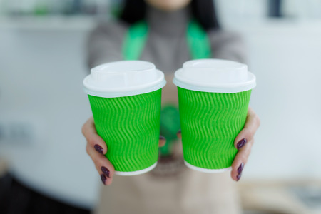 Barista in apron is holding in hands hot coffee in green takeaway paper cup. Coffee take away at...