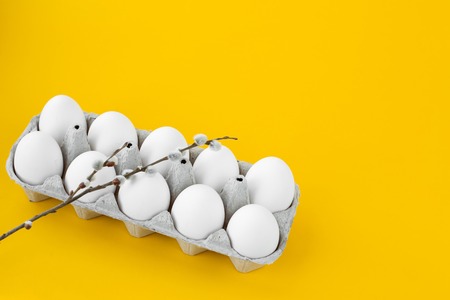 White chicken eggs in an open cardboard box on a yellow background. A willow twig lies on top....
