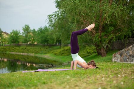 Slender young brunette yogi performs complex yoga exercise on green grass in the summer on a...