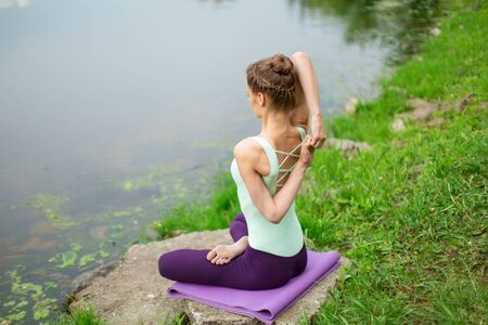 Young beautiful caucasian brunette girl doing yoga on a green lawn against the background of the...