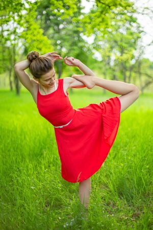 Slim beautiful brunette girl in a red dress performs yoga poses in a summer park. Green forest at sunset.の写真素材