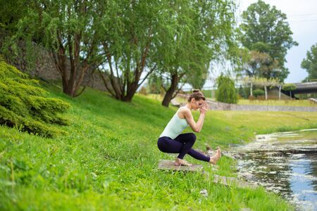 Young beautiful caucasian brunette girl doing yoga on a green lawn against the background of the river.の写真素材