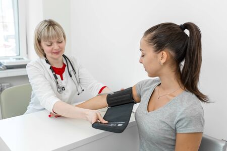 Professional female doctor putting on device for measuring pressure on young female patient hand in...