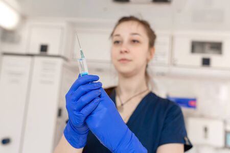 Young professional nurse in medical uniform, show syringe in her hands at ambulance background.