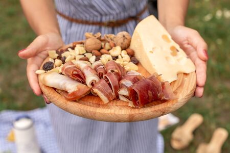 Young woman holds a wooden dish with delicacies. Snacks for a summer picnic jamon, cheese, dry fruits, jerky and nuts.の写真素材