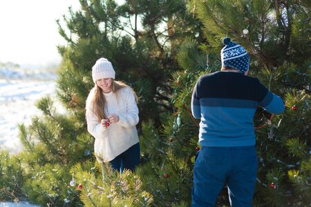a guy with a girl decorates a green Christmas tree on a street in the winter in the forest with decorative toys and garlands. Christmas tree decorations.の写真素材