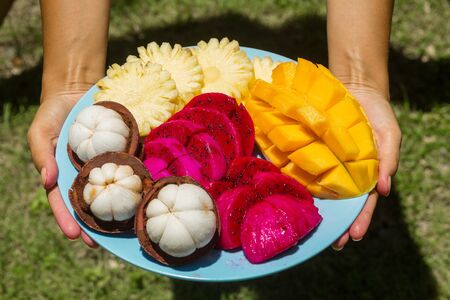 A girl in the green jungle is holding a plate with freshly cut and peeled tropical fruits.