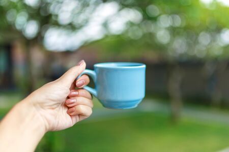 Closeup, girl holds in her hand a blue cup with a drink on a blurred background of a green yard.の写真素材