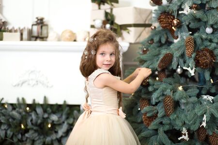A cute little girl in a lush beige dress hangs cones on the Christmas tree that is standing in the house. Christmas tale, happy childhood.の写真素材