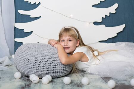 A little girl lies smiling on a pillow covered with a gray knitted blanket, against the background of an artificial white Christmas tree decor from cardboard, and next to it lies a garlandの写真素材