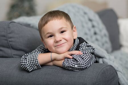 A little boy lies on a sofa covered with a gray knitted blanket and smiles, against the background...