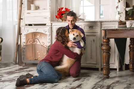Cheerful young couple is hugging and kissing a dog of Akita Inu breed. in Christmas decorations in retro style.の写真素材