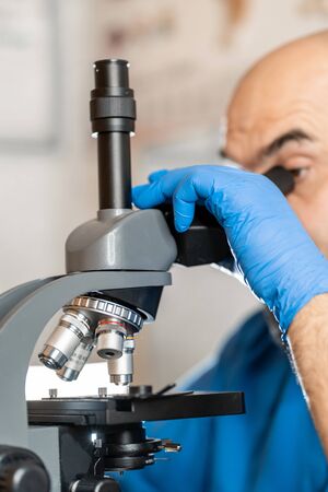 Male laboratory assistant examining biomaterial samples in a microscope. Cllose up hands in blue...