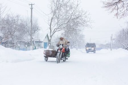 Winter, rural streets are covered with snow. A man rides a motorcycle in a snowstorm.の写真素材