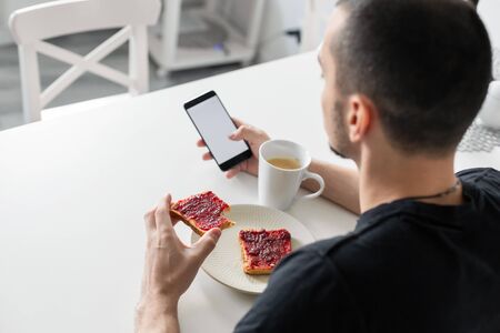 The guy watches the news on the phone during breakfast.