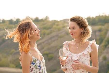 The company of gorgeous female friends having fun, drink wine, and enjoy hills landscape picnic.