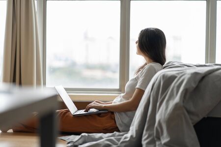 Beautiful young brunette girl working on a laptop, sitting on the floor near the bed by the...