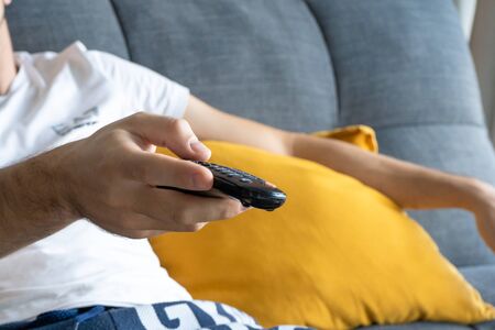 A thin guy in home clothes sits on a sofa and watches TV. Bored guy with tv remote control.の写真素材