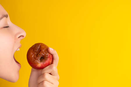 The girl bites a rotten apple with a worm on a yellow background. Expired products, junk food.の写真素材