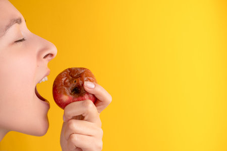 The girl bites a rotten apple with a worm on a yellow background. Expired products, junk food.の写真素材