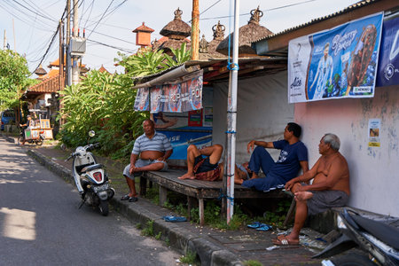 People resting near the road in an unusual Asian village. Children are playing and adults are talking sitting on the roadside. Bali, Indonesia - 11.30.2022のeditorial素材