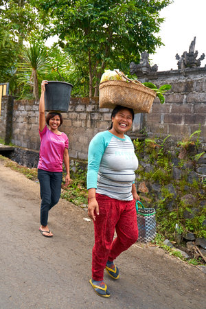 Local women carry baskets of food on their heads in an Indonesian village. Portrait of a smiling woman in Bali. Bali, Indonesia - 08/12/2022のeditorial素材
