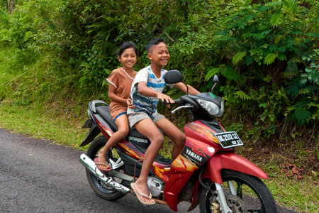 Children ride a scooter without helmets on a road in a village in Asia. Bali, Indonesia - 12.24.2022のeditorial素材
