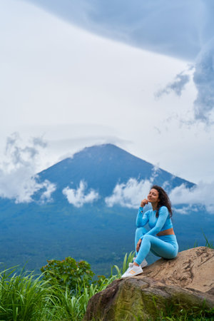A young woman sits on a large rock in a viewpoint and enjoys the view of the sacred Mount Agung volcano hidden by clouds on a rainy day on the island of Bali.の写真素材