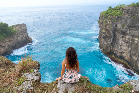 A beautiful woman in a pink dress sits on a cliff above the ocean on the island of Nusa Penida....