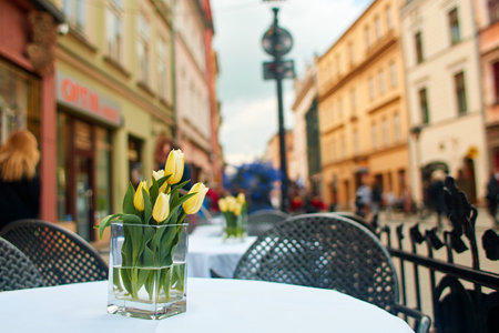 A romantic card with a bouquet of tulips. The spring yellow flowers are in a transparent glass vase on a table in a small cozy cafe on the old European streetsの写真素材