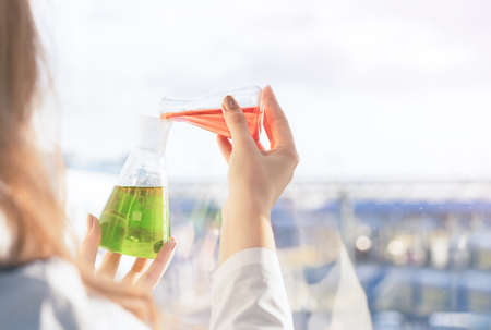 A girl laboratory assistant standing at the window mixes reagents in flasks in the laboratory at production. High quality photoの写真素材