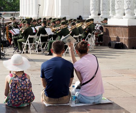A pre-adolescent girl and a pair of young men seated on the parapet during a performance of the brass band. Central Avenue in the Park of VDNH, Moscow. International Military Music Festival "Spasskaya Bashnya".のeditorial素材