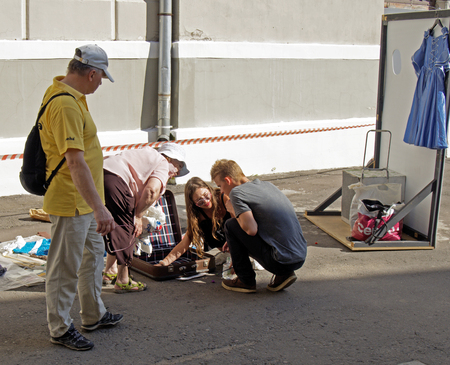 Squatting young adult girl and guy examine lying on the ground an open suitcase next to the bent over them by the seller senior adult woman at the flea market.のeditorial素材