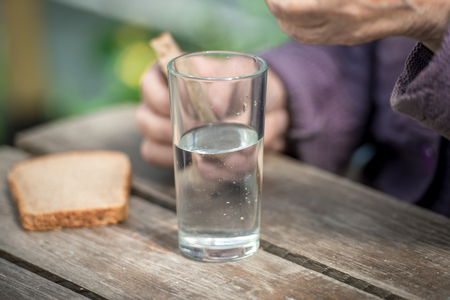 The old woman drinks water and eats bread at an old wooden table in a gardenの写真素材