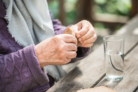 The old woman drinks water and eats bread at an old wooden table in a gardenの写真素材
