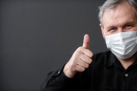 Happy man in a sterile face mask to escape from the covid-19 coronavirus during the quarantine pandemic, showing his thumb at the top on a dark backgroundの写真素材