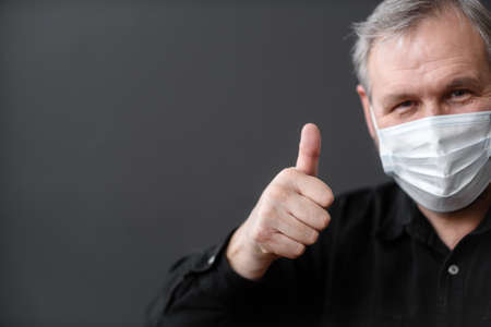 Happy man in a medical mask shows a thumbs up on a dark backgroundの写真素材