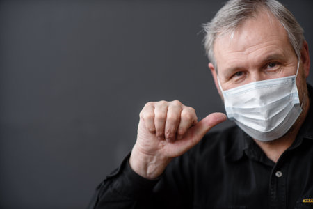 Happy man in a sterile face mask to escape from the covid-19 coronavirus during the quarantine pandemic, showing his thumb at the top on a dark backgroundの写真素材
