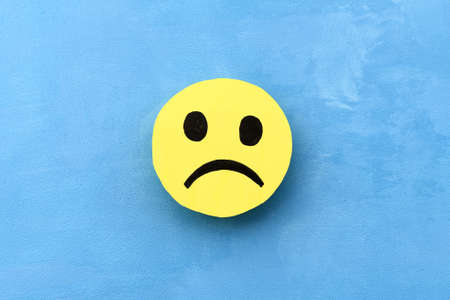 Yellow sad smiley face on a blue background. The concept of negative mood. Emotions and feedback.の写真素材