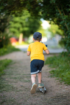 Little boy in a yellow T-shirt and a black cap riding a scooter in the parkの写真素材