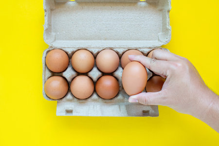 Man's hand is picking up a brown egg in a carton box. On a yellow background.