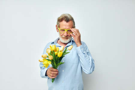 Senior grey-haired man in a blue shirt with a bouquet of flowers light backgroundの写真素材